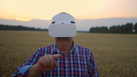 Senior-adult-farmer-in-a-virtual-reality-helmet-in-a-field-of-grain-crops.-In-the-sunset-light-an-elderly-man-in-a-tractor-driver-uses-virtual-reality-glasses.-VR-technologies-and-modern-agribusiness